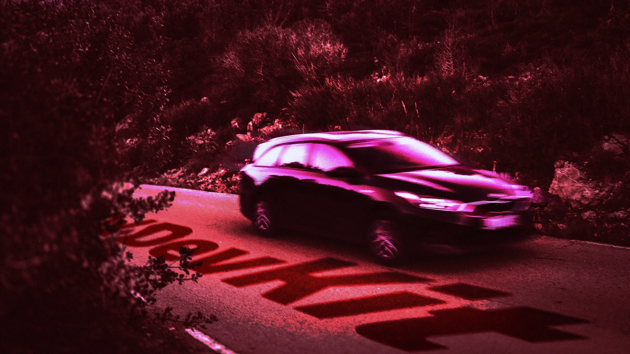 A dark car driving along a road with “10x DevKit” painted in large red block letters on the pavement. The scene appears to be at twilight, with dark, blurred foliage lining the roadside, all cast in shades of pink and deep maroon. The car, slightly blurred as if in motion, is dark and also tinged with pink, reflecting the overall color scheme of the image. The letters on the road seem almost projected or like a stencil, with a brighter area of pink around them contrasting against the darker road surface.